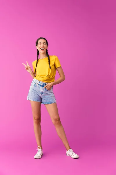 Pleine longueur photo de jeune femme joyeuse portant des shorts en denim clin d'oeil et signe de paix gestuel — Photo