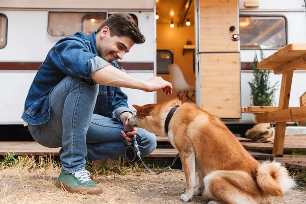 Portret van ongeschoren knappe man dragen denim kleren strelen rode hond terwijl kraken in de buurt van huis op wielen buitenshuis — Stockfoto