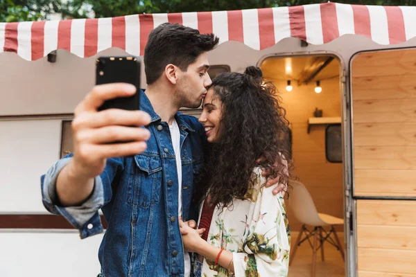 Εικόνα του ανθρώπου φιλιά όμορφη γυναίκα και τη λήψη selfie στο κινητό τηλέφωνο κοντά στο σπίτι σε τροχούς σε εξωτερικούς χώρους — Φωτογραφία Αρχείου