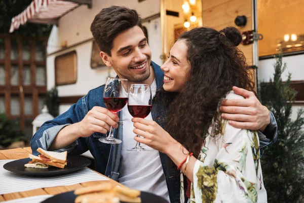 Retrato de casal sorrindo bebendo vinho tinto enquanto abraçando juntos na mesa de madeira perto de reboque ao ar livre — Fotografia de Stock