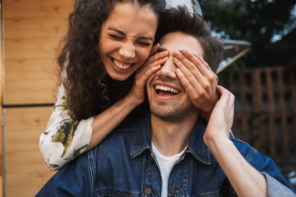 Porträt einer brünetten, lachenden Frau, die die Augen ihres Freundes in der Nähe des Wohnwagens im Freien verdeckt — Stockfoto