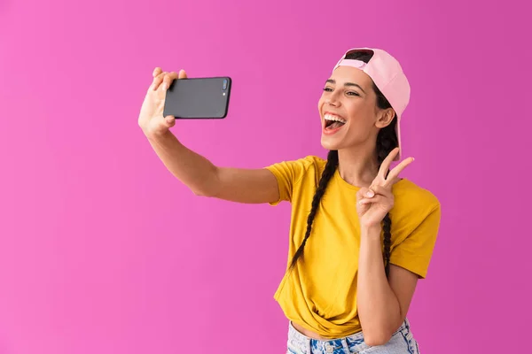 Image of joyous woman wearing cap showing peace sign and taking selfie photo on cellphone — Stock Photo, Image