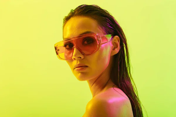 Incroyable jeune femme posant isolé sur fond de mur jaune avec néon lumières lumineuses portant des lunettes de soleil de mode . — Photo