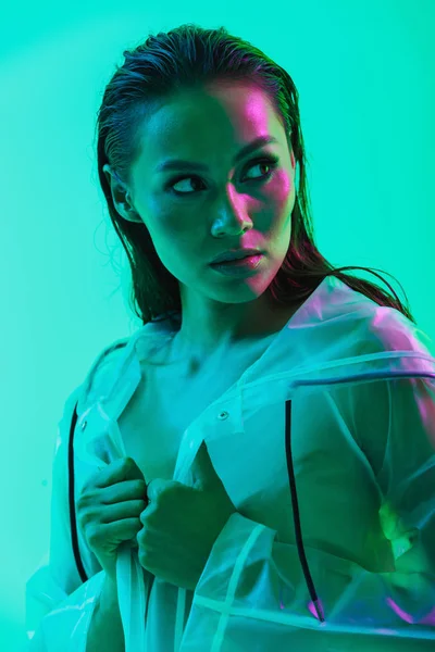 Mujer seria posando aislada sobre fondo de pared azul con luces brillantes de neón vestidas con impermeable . — Foto de Stock