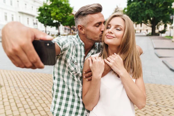 Bild eines fröhlichen jungen Paares, das sich küsst und zusammen Selfie-Fotos macht, während es durch die Straßen der Stadt läuft — Stockfoto