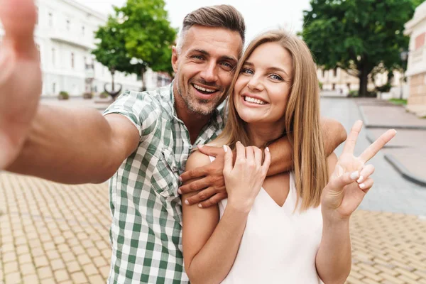 Bild eines hübschen jungen Paares, das ein Friedenszeichen zeigt und zusammen ein Selfie-Foto macht, während es durch die Straße geht — Stockfoto