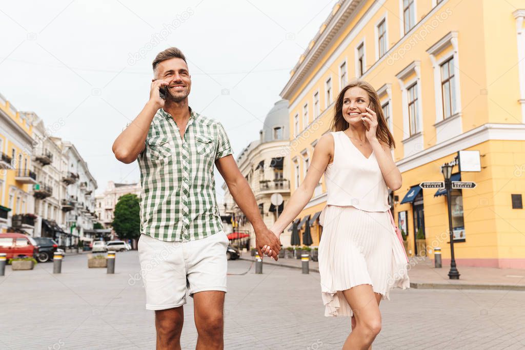 Image of happy young couple holding hands together and talking on smartphones while walking through city street