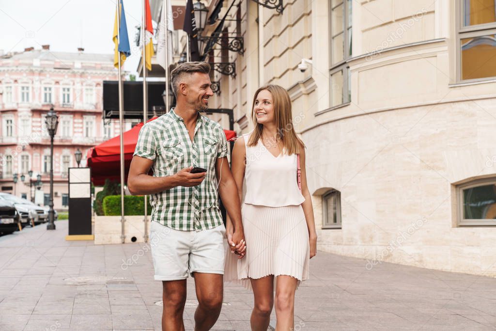 Image of pleased young couple listening music together with earphones while walking through city street