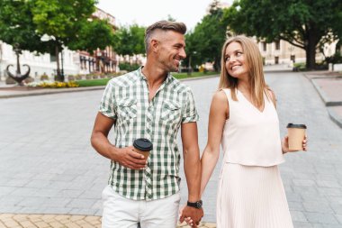 Kağıt bardaklarla şehir caddesinde yürürken el ele tutuşan büyüleyici genç çiftin görüntüsü