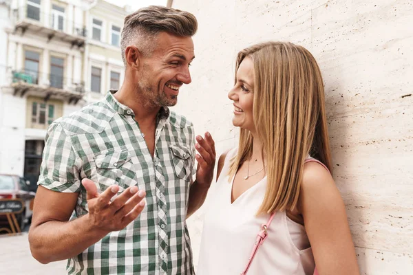 Bild eines aufgeregten blonden Paares in Sommerkleidung, das zusammen lächelt und spricht, während es an der Stadtstraße gegen eine Mauer steht — Stockfoto