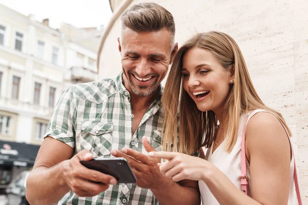 Imagem do alegre casal animado sorrindo e olhando para o celular juntos enquanto estão de pé contra a parede na rua da cidade — Fotografia de Stock