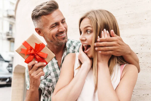 Bild eines erstaunten aufgeregten Paares, das lächelt und die Geschenkbox zusammenhält, während es an der Stadtstraße gegen eine Mauer steht — Stockfoto