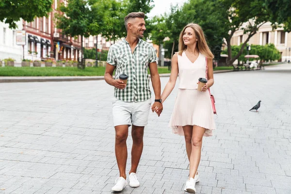 Bild eines glücklichen jungen Paares, das Händchen hält, während es mit Pappbechern durch die Stadt läuft — Stockfoto