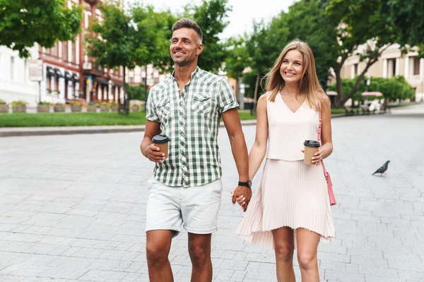 Kép a örömteli fiatal pár kezében együtt sétálva a városi utcán papírpoharak — Stock Fotó