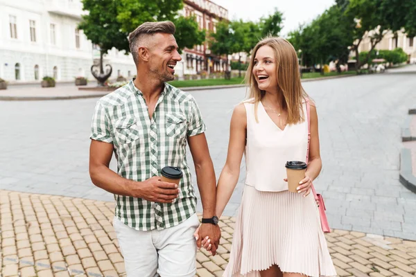 Kép a vidám fiatal házaspár kezében együtt séta a városi utcában papírpoharak — Stock Fotó