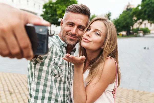 Obraz bělošského mladého páru, který se usmívá a při chůzi po městské ulici si bere fotografii. — Stock fotografie