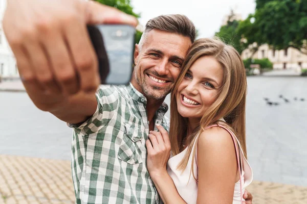 Bild eines zufriedenen jungen Paares, das lächelt und zusammen Selfie-Fotos macht, während es durch die Straßen der Stadt läuft — Stockfoto