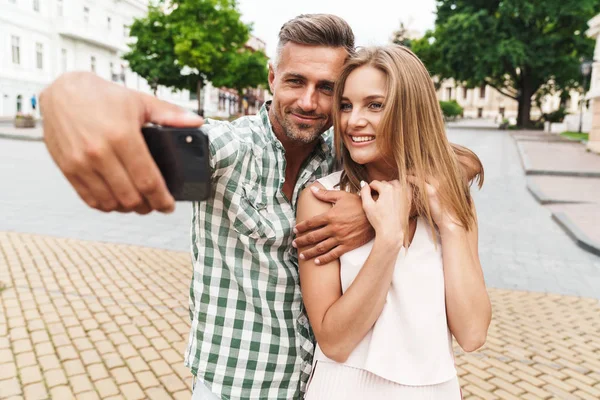 Immagine di una giovane coppia felice che sorride e scatta foto di selfie insieme mentre cammina per strada — Foto Stock