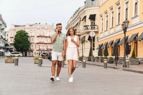 Imagem de belo casal feliz de mãos dadas e usando smartphones enquanto caminha pela rua da cidade — Fotografia de Stock