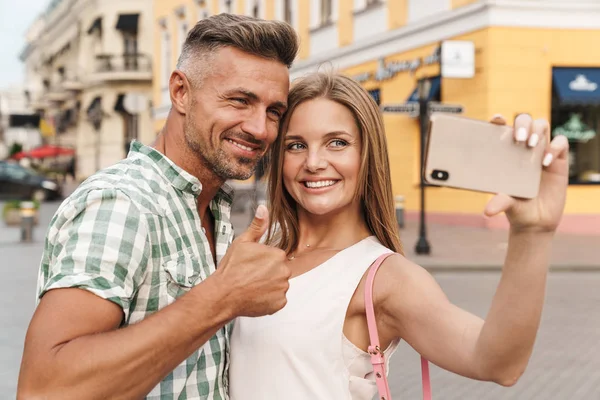 Foto de jovem casal feliz sorrindo e mostrando o polegar para cima ao tirar foto selfie na rua da cidade — Fotografia de Stock
