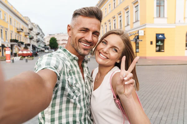 Foto de amor jovem casal sorrindo e mostrando sinal de paz ao tirar foto selfie na rua da cidade — Fotografia de Stock