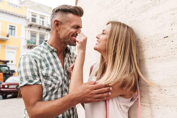 Bild eines schönen blonden Paares, das zusammen lächelt und flirtet, während es auf der Straße der Stadt gegen eine Mauer steht — Stockfoto