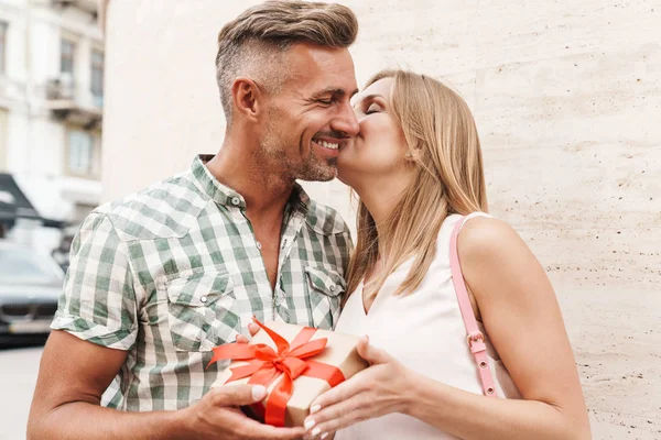 Bild eines ziemlich aufgeregten Paares, das sich küsst und Geschenke zusammenhält, während es an der Stadtstraße gegen eine Mauer steht — Stockfoto