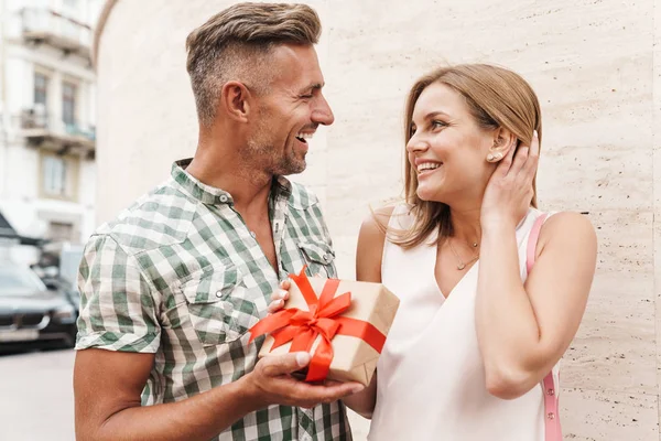 Bild eines romantischen aufgeregten Paares, das lächelt und die Geschenkbox zusammenhält, während es an der Stadtstraße gegen eine Mauer steht — Stockfoto