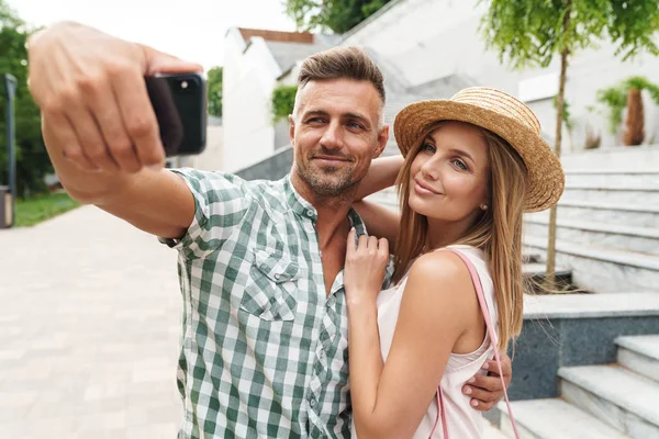Foto de jovem casal caucasiano sorrindo e abraçando juntos enquanto tirando foto selfie na rua da cidade — Fotografia de Stock