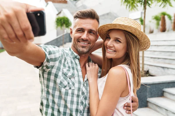 Foto de casal feliz caucasiano sorrindo e abraçando juntos enquanto tirando foto selfie na rua da cidade — Fotografia de Stock