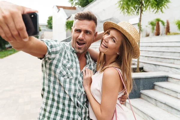 Photo of attractive caucasian couple smiling and hugging together while taking selfie photo on city street — Stock Photo, Image