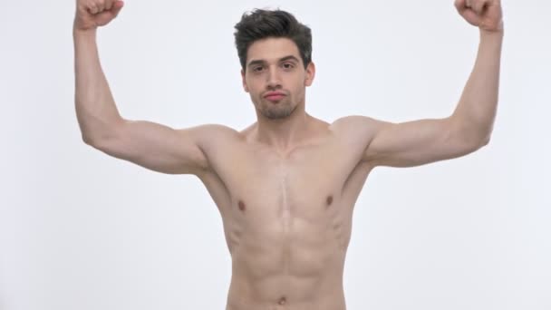 白い背景の上にカメラを見ながら 裸の胴子を持つ自信のある若いブルネットの男は 腕を上げて彼の二頭筋を示しています — ストック動画