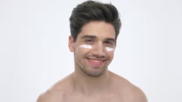 白い背景の上にカメラを見ながら 裸の胴子が目のしわにクリームで微笑む陽気な若いブルネットの男のクローズアップビュー — ストック動画