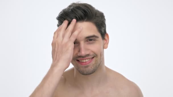 白い背景の上にカメラを見ながら 裸の胴子の笑顔と手で髪を固定する幸せな若いブルネットの男のクローズアップビュー — ストック動画