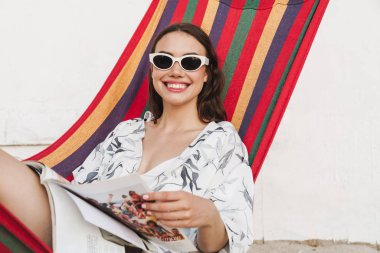 Olumlu memnun bir hamak okuma dergisi üzerinde poz sahilde gülümseyen genç güzel kadın.