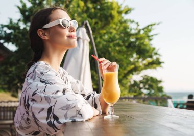 Neşeli mutlu memnun genç güzel kadın plajda güneş gözlüğü takan kafede meyve suyu kokteyli içme.