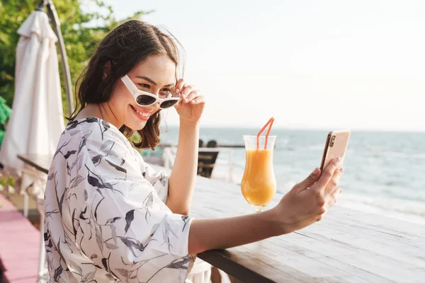 Linda joven feliz hermosa mujer en la playa bebiendo jugo cóctel en la cafetería con gafas de sol tomar una selfie por teléfono . — Foto de Stock