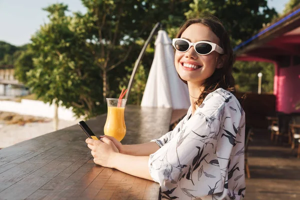 Sorridente carino allegra giovane bella donna in spiaggia bere succo di cocktail in caffè indossando occhiali da sole utilizzando il telefono cellulare . — Foto Stock