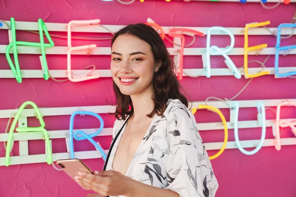 Mujer bonita joven positiva en la fiesta de la playa con brillante led fondo colorido utilizando el teléfono móvil . —  Fotos de Stock