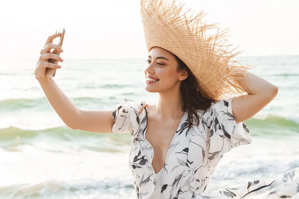 아름다운 화창한 아침에 걷는 해변에서 행복한 젊은 아름다운 여성이 휴대 전화로 셀카를 찍습니다.. — 스톡 사진