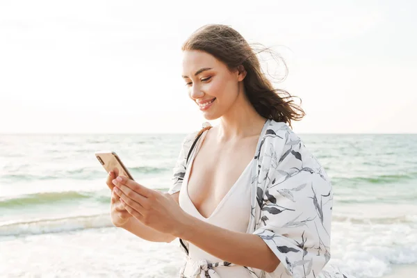 Sonriendo feliz feliz joven hermosa mujer en la playa caminando en una hermosa mañana soleada utilizando el teléfono móvil . — Foto de Stock