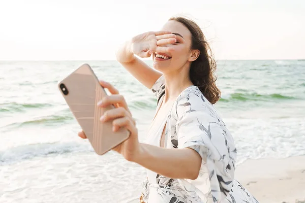 Plăcut fericit tânără frumoasă femeie la plaja de mers pe jos într-o dimineață frumoasă însorită ia un selfie de telefon mobil . — Fotografie, imagine de stoc