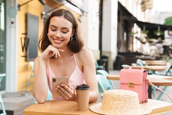 Image d'une femme brune heureuse souriante et tenant son téléphone portable alors qu'elle était assise dans un café d'été dans la rue — Photo