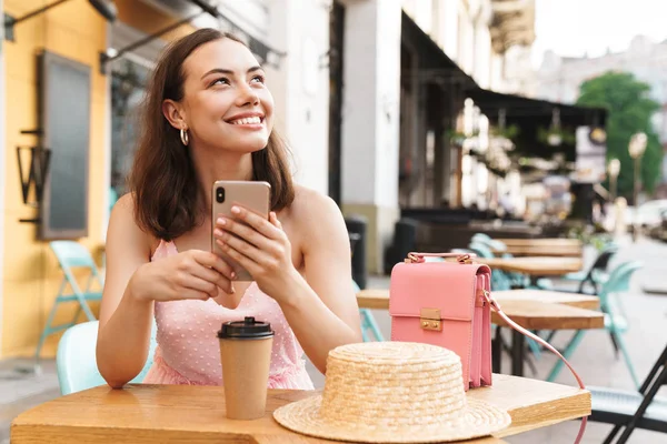 Image d'une femme brune heureuse souriante et tenant son téléphone portable alors qu'elle était assise dans un café d'été dans la rue — Photo