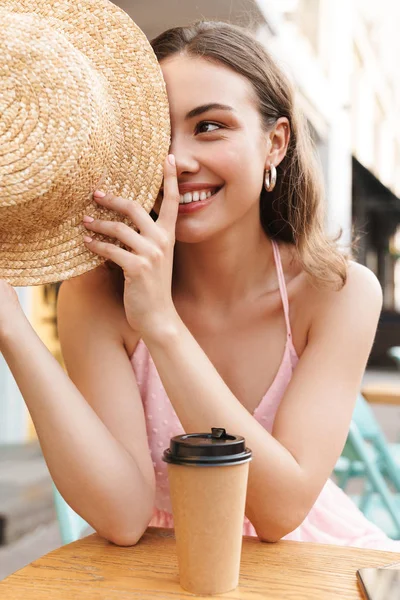 Imagen de la linda mujer joven sonriendo y sosteniendo sombrero de paja mientras está sentado en la calle café de verano —  Fotos de Stock