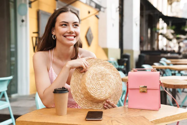Изображение привлекательной брюнетки, улыбающейся и держащей соломенную шляпу, сидя в уличном летнем кафе — стоковое фото