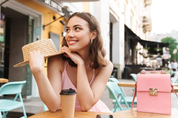 Immagine di donna bruna glamour che sorride e tiene il cappello di paglia mentre si siede in un caffè estivo di strada — Foto Stock