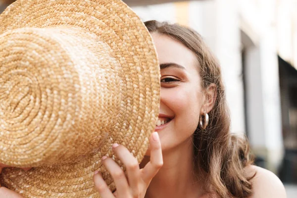 Immagine di una bella donna bruna che sorride e tiene il cappello di paglia mentre è seduta in un caffè estivo di strada — Foto Stock