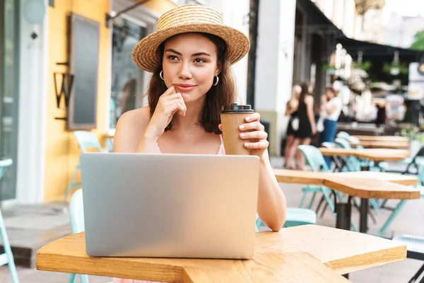 Imagen de la mujer morena relajada sonriendo y utilizando el ordenador portátil mientras está sentado en la calle café de verano — Foto de Stock