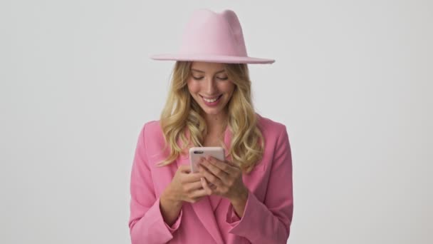 ピンクのジャケットと帽子の陽気な若いブロンドの女性は 孤立した灰色の背景の上にスマートフォンを使用しながら笑顔と笑い — ストック動画